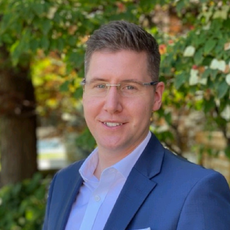 Headshot of Geoff Ramsay - Blue suit, glasses, white shirt, blurred trees in background